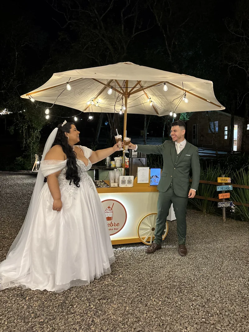 Noivos brindando com milkshakes diante do carrinho iluminado da Celebre em um casamento ao ar livre à noite